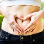 A 25 year old caucasian woman with fair skin making a heart shape with her hands over her stomach, in a garden