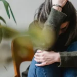 Stressed young woman holding her head surrounded by green plants, symbolizing anxiety and natural healing